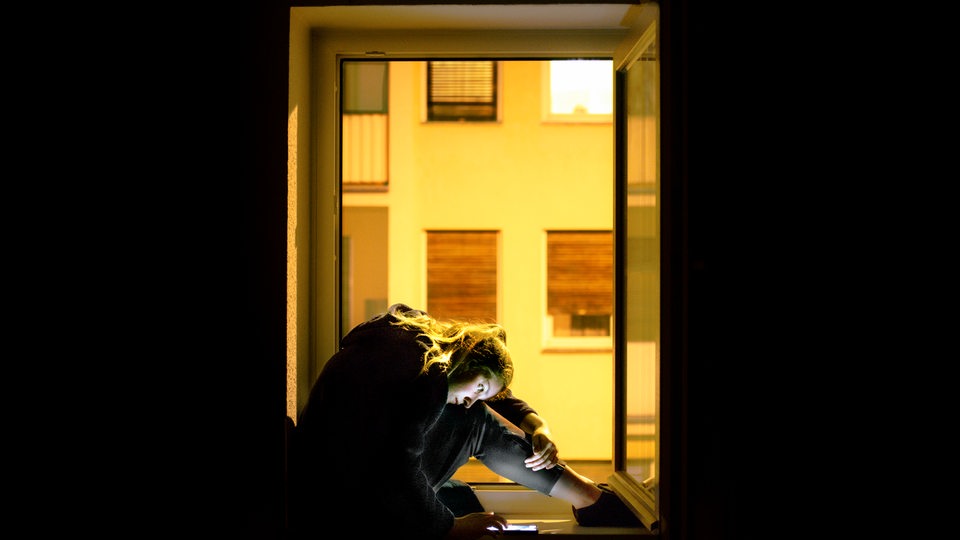 Eine Frau sitzt vor einem Fenster. Im Hintergrund sieht man in gelbes Licht getaucht die Fenster eines gegenüberliegenden Wohnhauses. Die auf dem Fenstersims sitzende Frau hat die Beine angewinkelt und schaut runter auf ihr Handy.