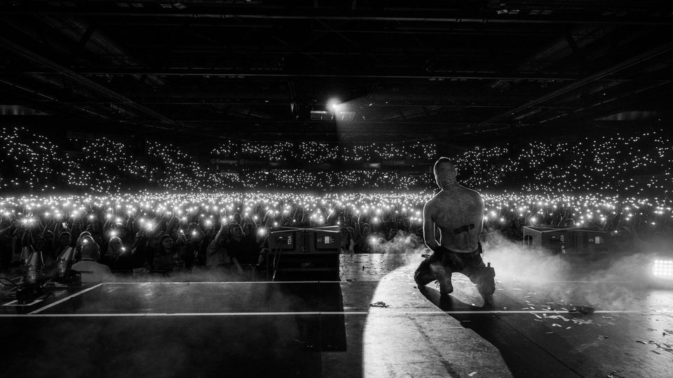 Schwarzweiß Bild, auf dem man Kontra K auf einer Bühne kniend sieht. Im Hintergrund eine Flut aus Lichtern seiner Fans.