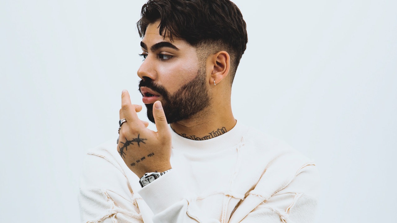 Rapper Monet192 in weiß gekleidet vor weißem Hintergrund. Er schaut zur Seite, seine Arme verschränkt, eine Hand unters Kinn angehoben. 