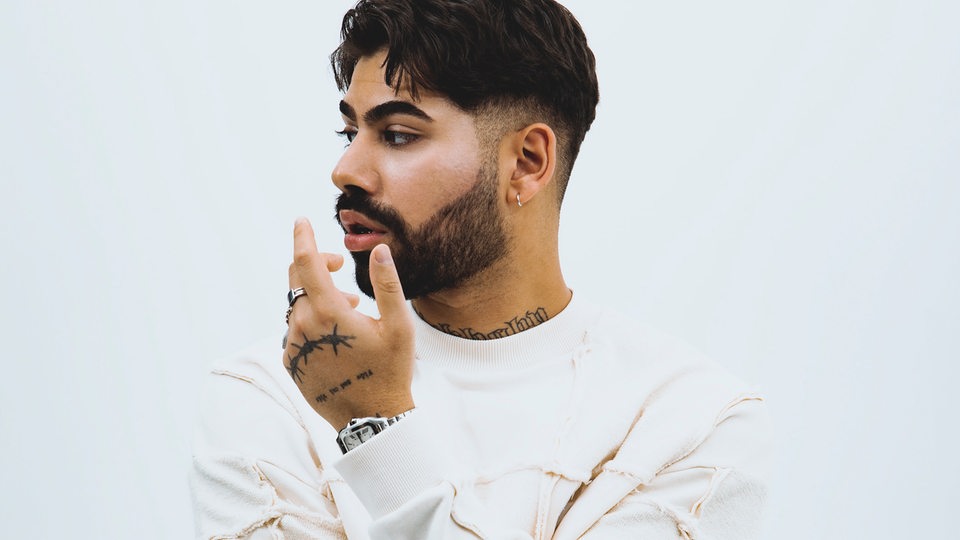 Rapper Monet192 in weiß gekleidet vor weißem Hintergrund. Er schaut zur Seite, seine Arme verschränkt, eine Hand unters Kinn angehoben. 