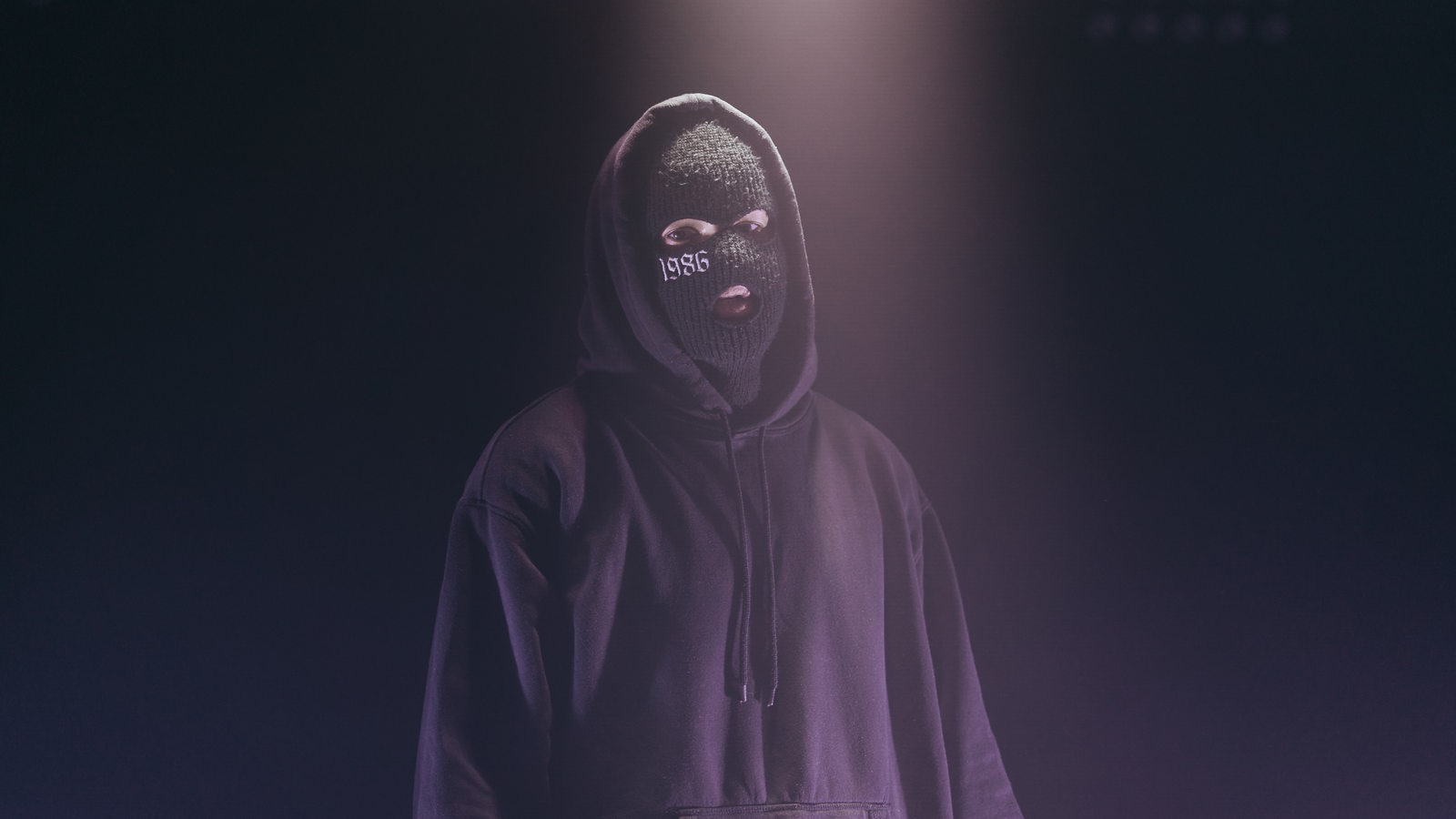 Der Künstler steht mit seiner Sturmmaske im Kapuzenhoodie vor der Kamera. Der Hintergrund ist dunkles lila, er wird von oben mit Licht sanft angestrahlt und blickt durch die Maske hindurch in die Kamera.