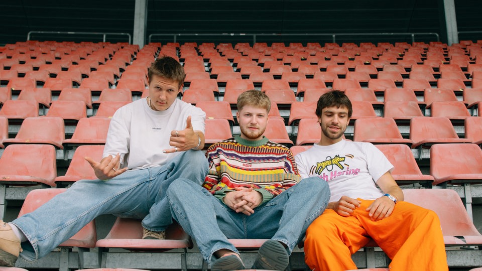Die Künstler der Band 01099 Gustav, Paul und Zachi sitzen auf roten Stühlen, vermutlich in einem Stadium und blicken in Richtung Kamera.