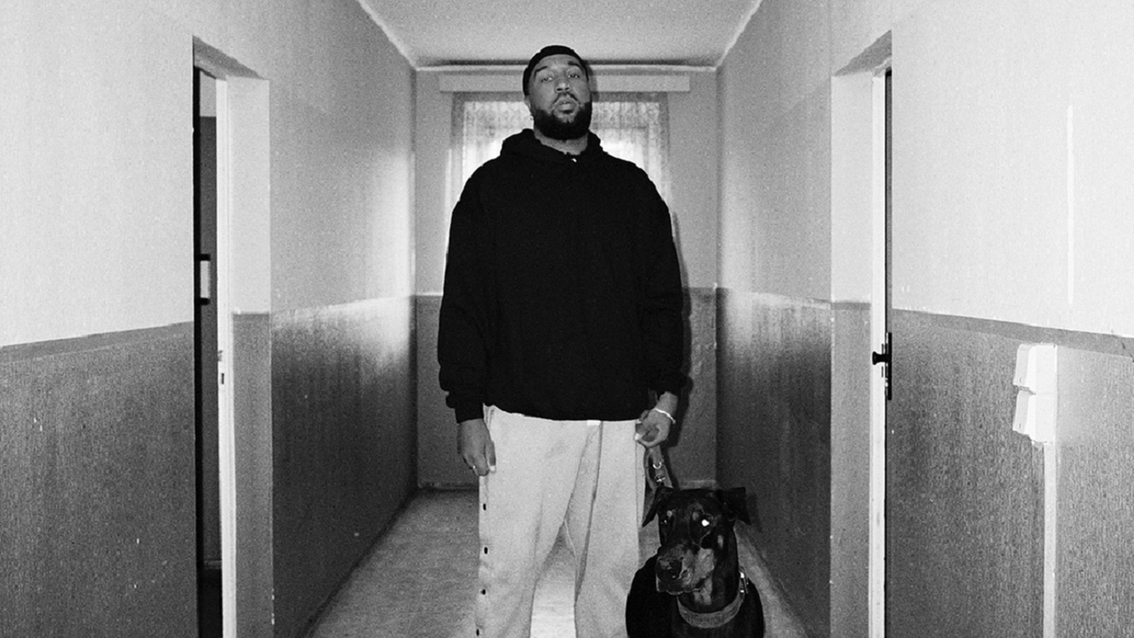 Ein schwarzweiß gegradetes Bild von OG Keemo, auf dem er mit einem Hund zu sehen ist. Er steht in einem Gang, links und rechts befinden sich geschlossene Türen.