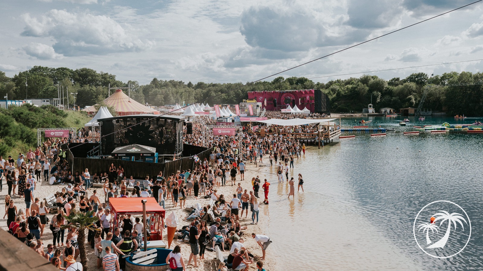 Das Bild zeigt ein Open-Air Festival mit einem Strand auf dem links eine Festivalbühne aufgebaut ist. Davor sammeln sich viele Menschen, die feiern. Rechts geht der Strand ins Wasser über, im hinteren Bereich schwimmende Menschen. Im Hintergrund ist eine Baumreihe am Horizont zu erkennen, davor eine weitere Bühne.