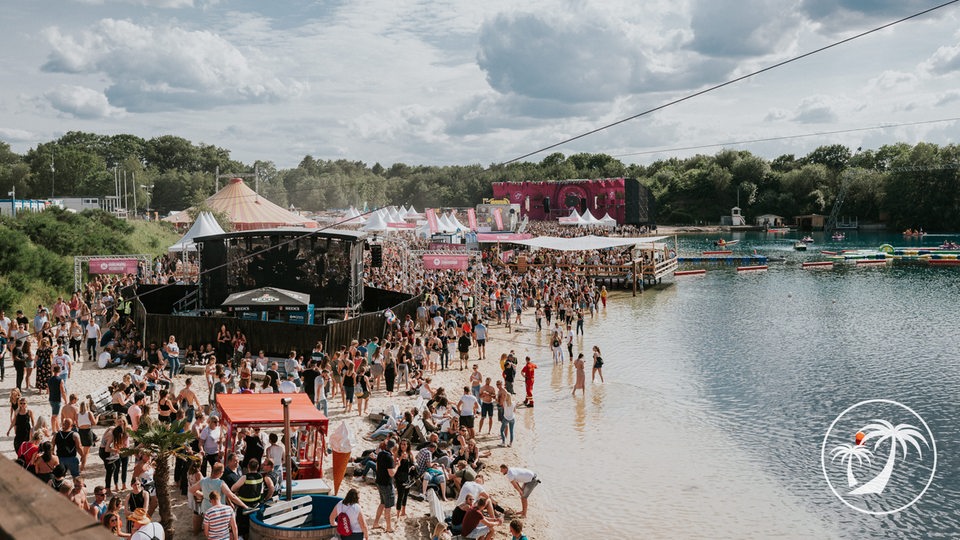 Das Bild zeigt ein Open-Air Festival mit einem Strand auf dem links eine Festivalbühne aufgebaut ist. Davor sammeln sich viele Menschen, die feiern. Rechts geht der Strand ins Wasser über, im hinteren Bereich schwimmende Menschen. Im Hintergrund ist eine Baumreihe am Horizont zu erkennen, davor eine weitere Bühne.
