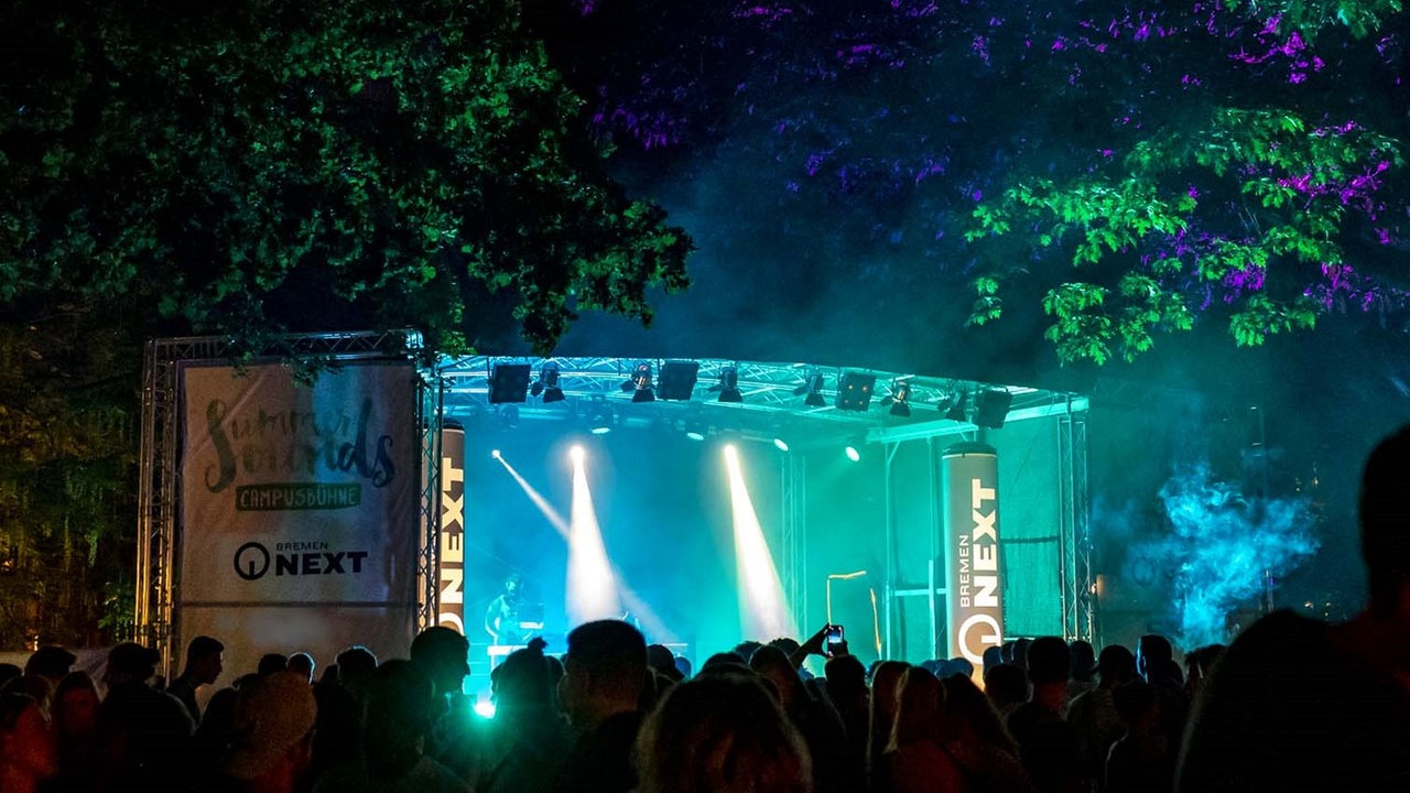 Man sieht die Bremen NEXT Bühne des Summer Sound Festivals. Es ist größtenweils dunkel, doch blau-grüne Neonlichter erhellen Bühne und umzu. Vor der Bühne eine Menschenmenge.