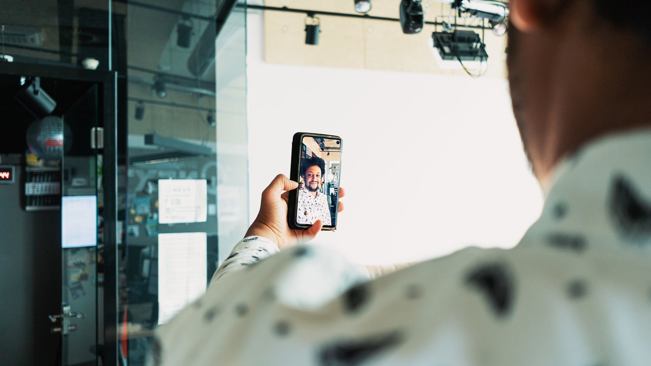 Eine junger Mann zeigt mit einem Handy per Videostream, wie er in einem Hörfunkstudio arbeitet.