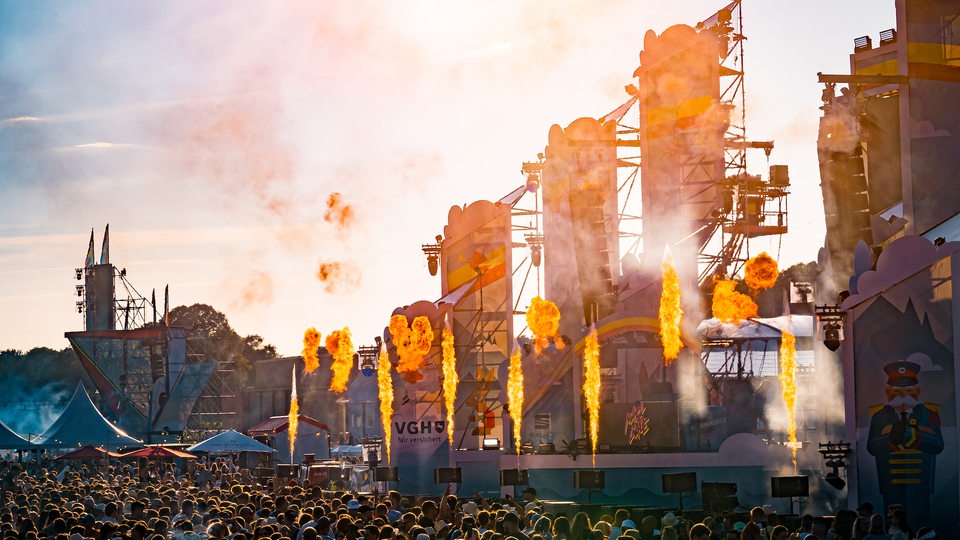 Bühne des Festivals mit mehreren Flammenwerfern, davor eine Menge feiernder Menschen.