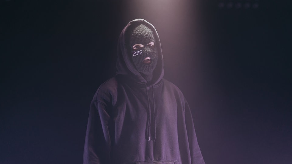 Der Künstler steht mit seiner Sturmmaske im Kapuzenhoodie vor der Kamera. Der Hintergrund ist dunkles lila, er wird von oben mit Licht sanft angestrahlt und blickt durch die Maske hindurch in die Kamera.