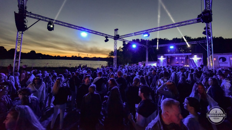 Das Bild zeigt eine Menge tanzender Menschen vor einer aufgebauten Bühne (hinten rechts) und dem Unisee (hinten links) bei Sonnenuntergang. Angestrahlt werden sie von blauen Leuchtstrahlern.