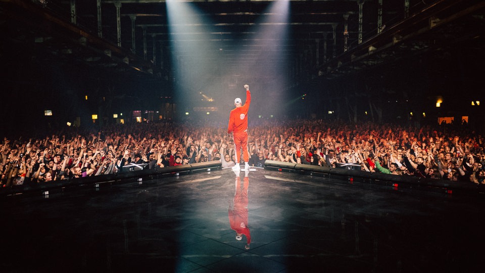 Sido steht mit dem Rücken zum Fotographen in einem roten Outfit am Rand einer Bühne. Vor ihm eine jubelnde Menschenmasse in einem dunkel abgelichteten Konzertraum.