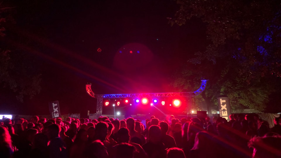 Das Bild zeigt die Uni Nacht Bühne aus 2022. Es ist dunkel, die Menschenmenge wird von pink-rotem Licht angestrahlt.